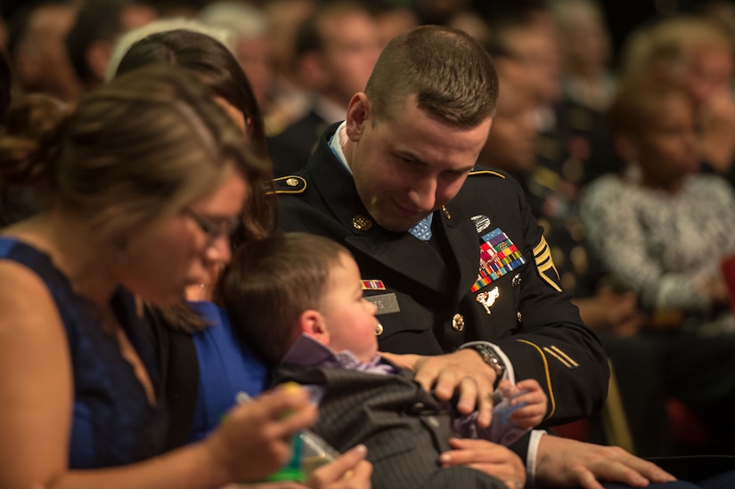A soldier in a seated crowd puts his hand on his small son’s chest.