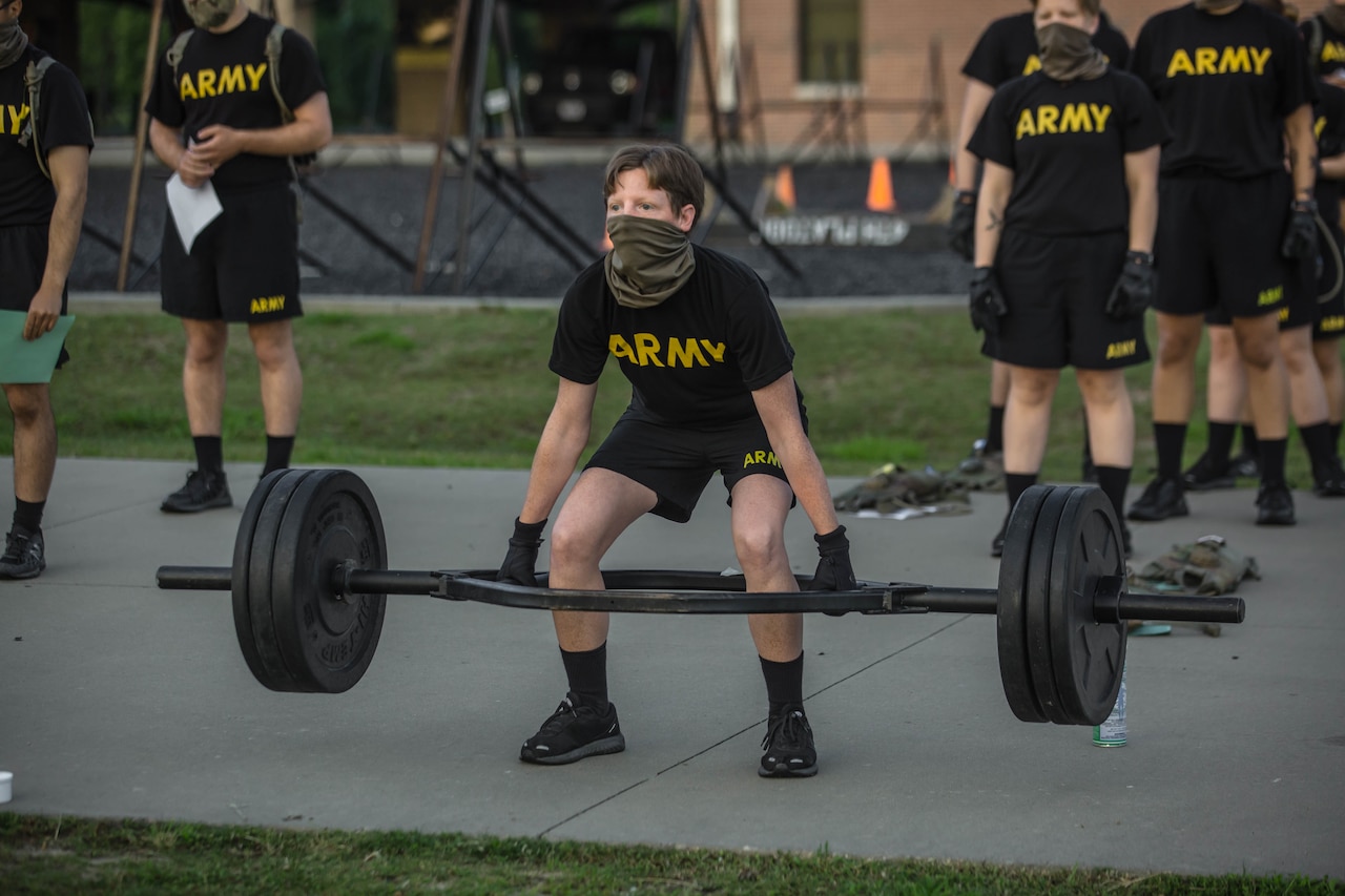 A service member lifts a barbell; others wait nearby.