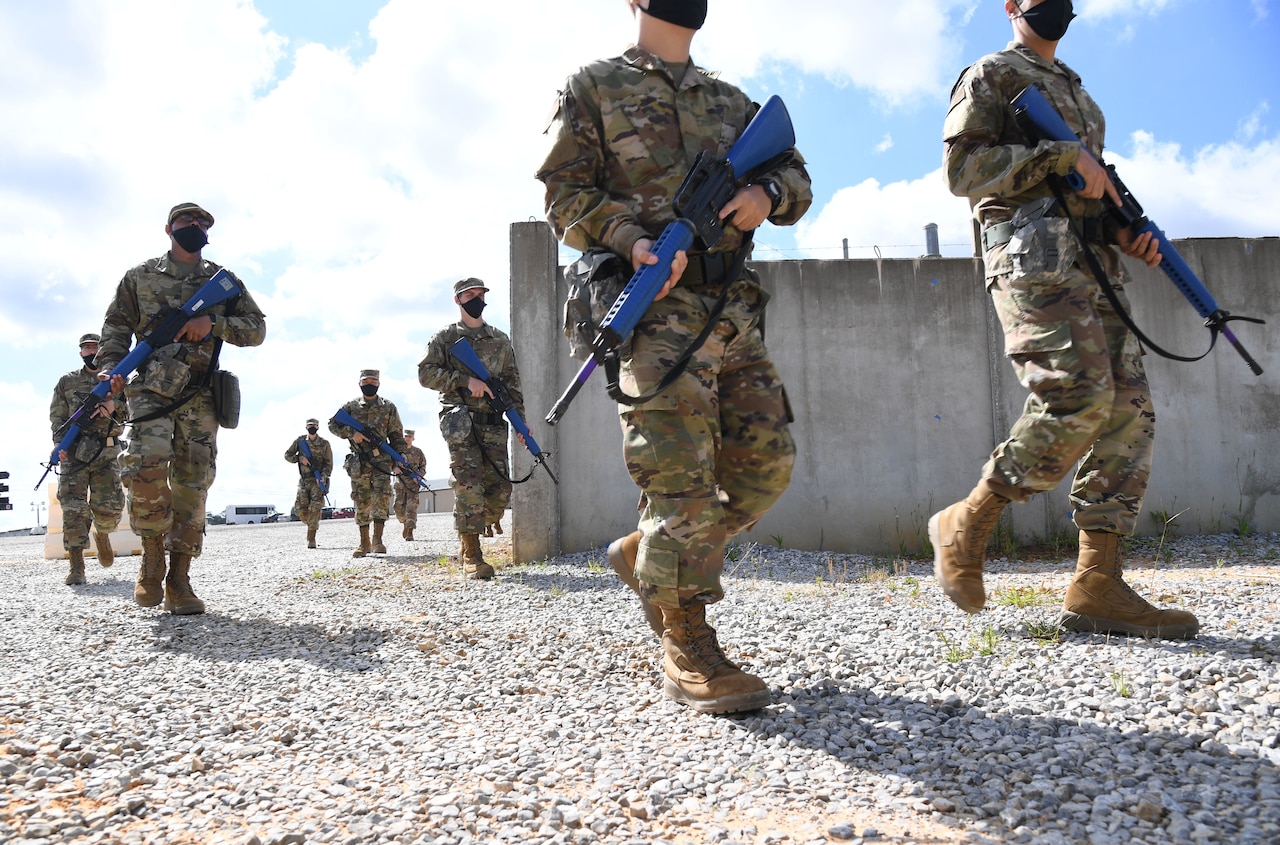 Service members walk on gravel carrying blue training rifles.