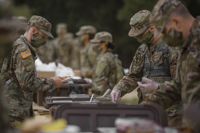 Service members wearing face masks gather around an outdoor table with portable meal containers.
