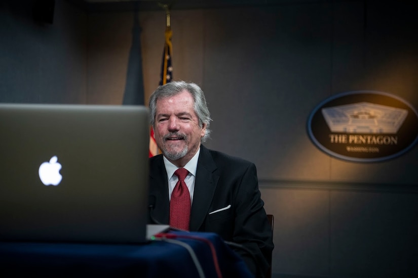 A man sits at a computer while a Pentagon sign hangs behind him.