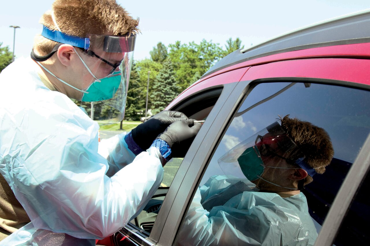 A soldier wearing protective gear administers a COVID-19 test to a motorist through a car’s open window.