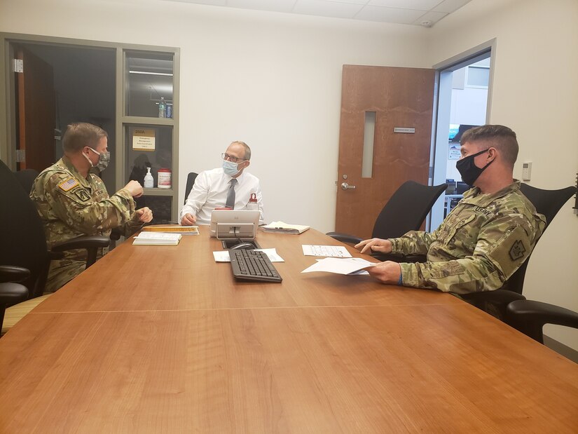 Two soldiers and a civilian, all wearing face masks, talk as they sit at a conference table.