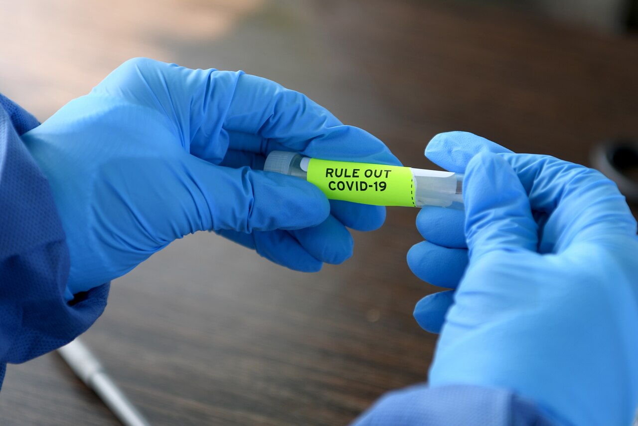An airman wearing protective equipment holds a vial containing a COVID-19 sample.