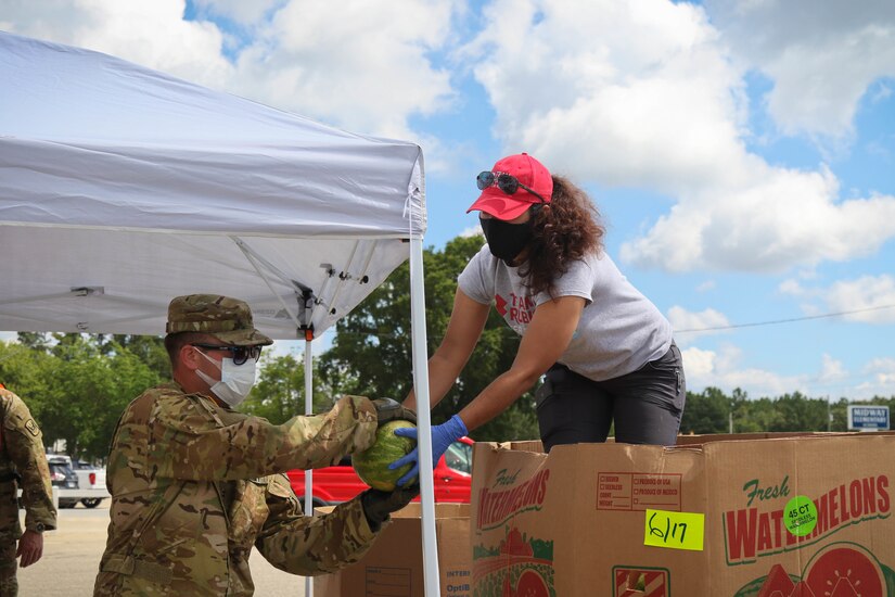 A soldier hands a watermelon to a woman in the bed of a truck. Both are wearing face masks.