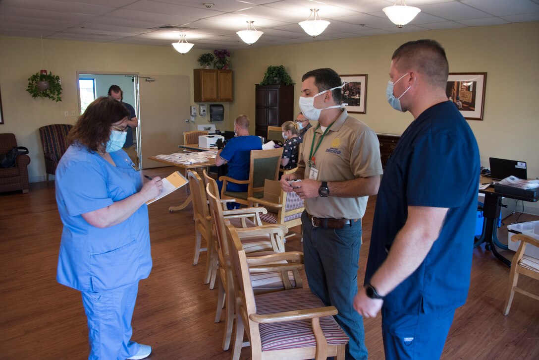 A nurse holding a clipboard speaks with two men. All are wearing face masks.