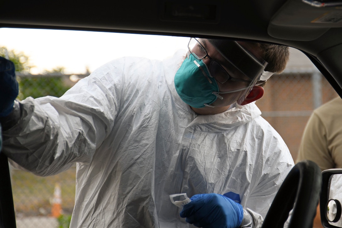 A guardsman in protective gear performs a nasal swab through a car window.