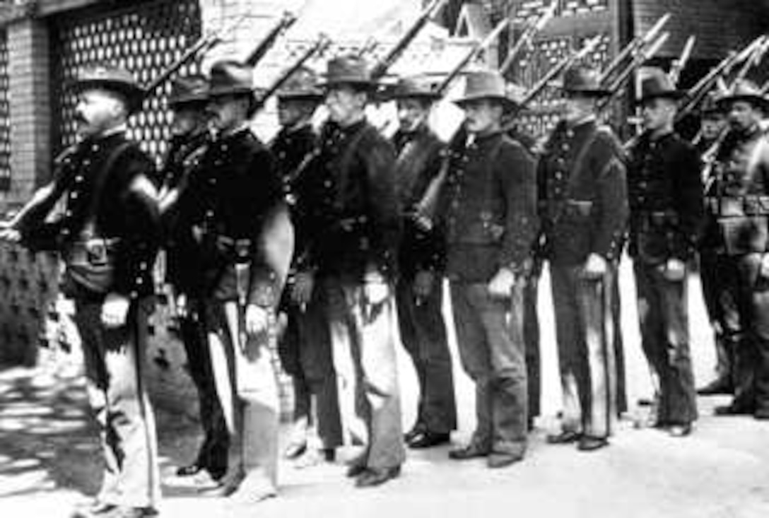 Men with rifles over their shoulders stand in line and at attention.