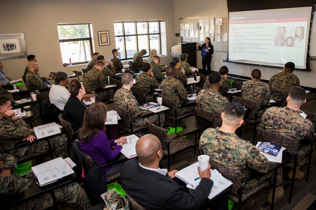 A group of people, some in military uniform, sit in a room watching a presentation on screen.