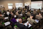 A group of people, some in military uniform, sit in a room watching a presentation on screen.