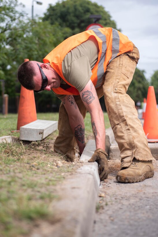 Staff Sgt. Jeffrey Elwell, 100th Civil Engineer Squadron heavy equipment horizontal engineer supervisor, positions a curb into place at RAF Mildenhall, England, July 12, 2020. Members of the pavement and construction equipment section, known as “Dirt Boyz,” removed unnecessary vehicle barriers to improve traffic flow. (U.S. Air Force photo by Airman 1st Class Joseph Barron)
