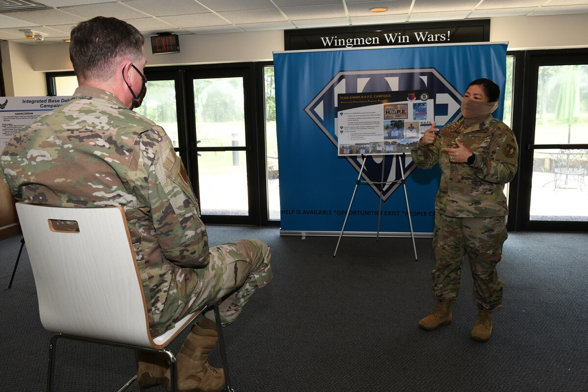 Photo shows one individual sitting in a chair listening to a briefing given by another individual in front of an easel and banner.
