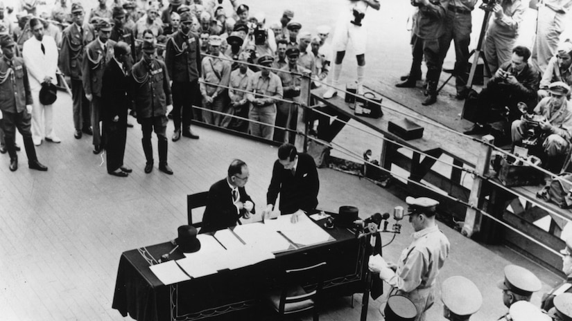A man sits behind a desk covered with papers.
