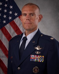 Official photo of Air Force officer in uniform seated in front of the United States of America flag.