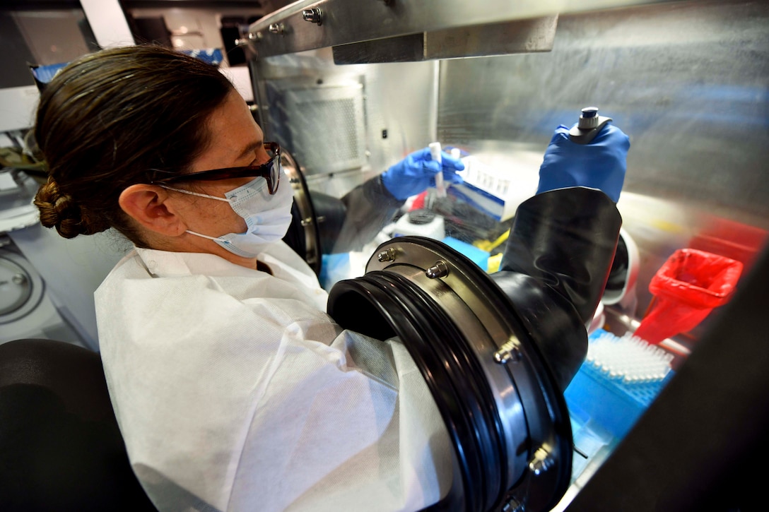 A soldier wearing protective gear looks at test tubes through a glass barrier.