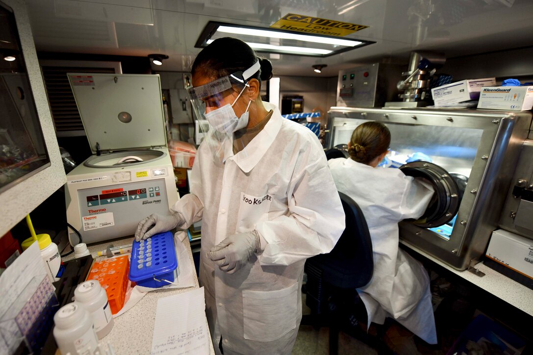 A soldier in protective gear touches test tubes and a service member works in the background.