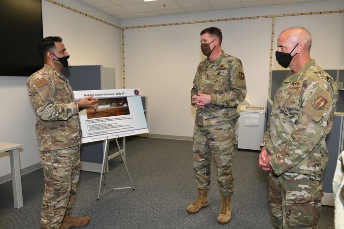 Photo shows three individual standing and speaking to each other in front of an easel.