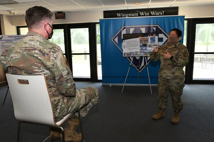 Photo shows one individual sitting in a chair listening to a briefing given by another individual in front of an easel and banner.