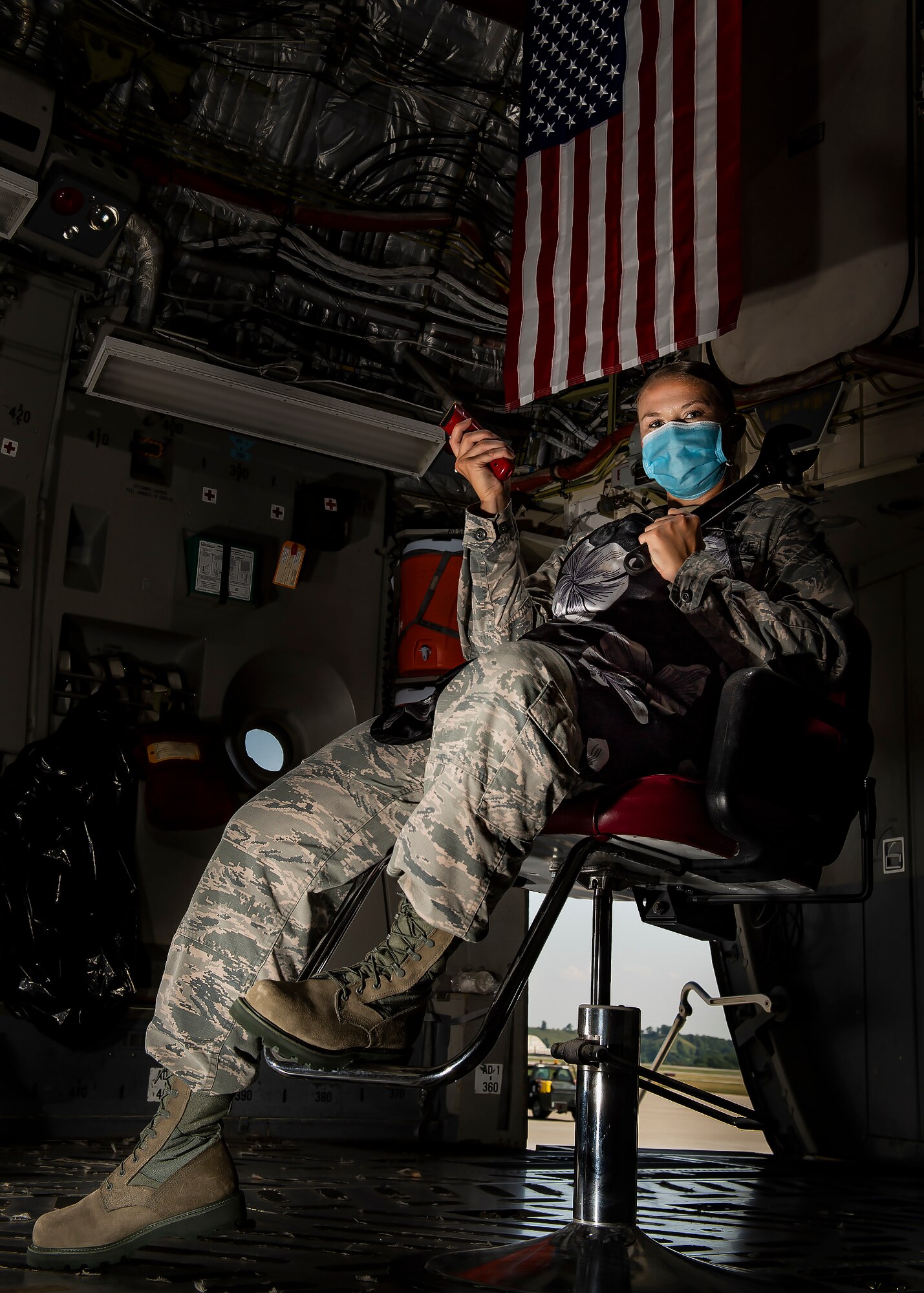 Senior Airman Sara Gutherie, 911th Aircraft Maintenance Squadron integrated flight control systems mechanic, poses for a photo inside a C-17 Globemaster III with a wrench and hair clippers while wearing an apron and personal protective equipment at the Pittsburgh International Airport Air Reserve Station, Pennsylvania, July 8, 2020. During COVID-19 restrictions, Gutherie helped her fellow Airmen by using her skills as a licensed cosmetologist to give them haircuts to ensure they stayed within regulations while taking donations for the Needy Airman Fund in return. (U.S. Air Force photo by Joshua J. Seybert)