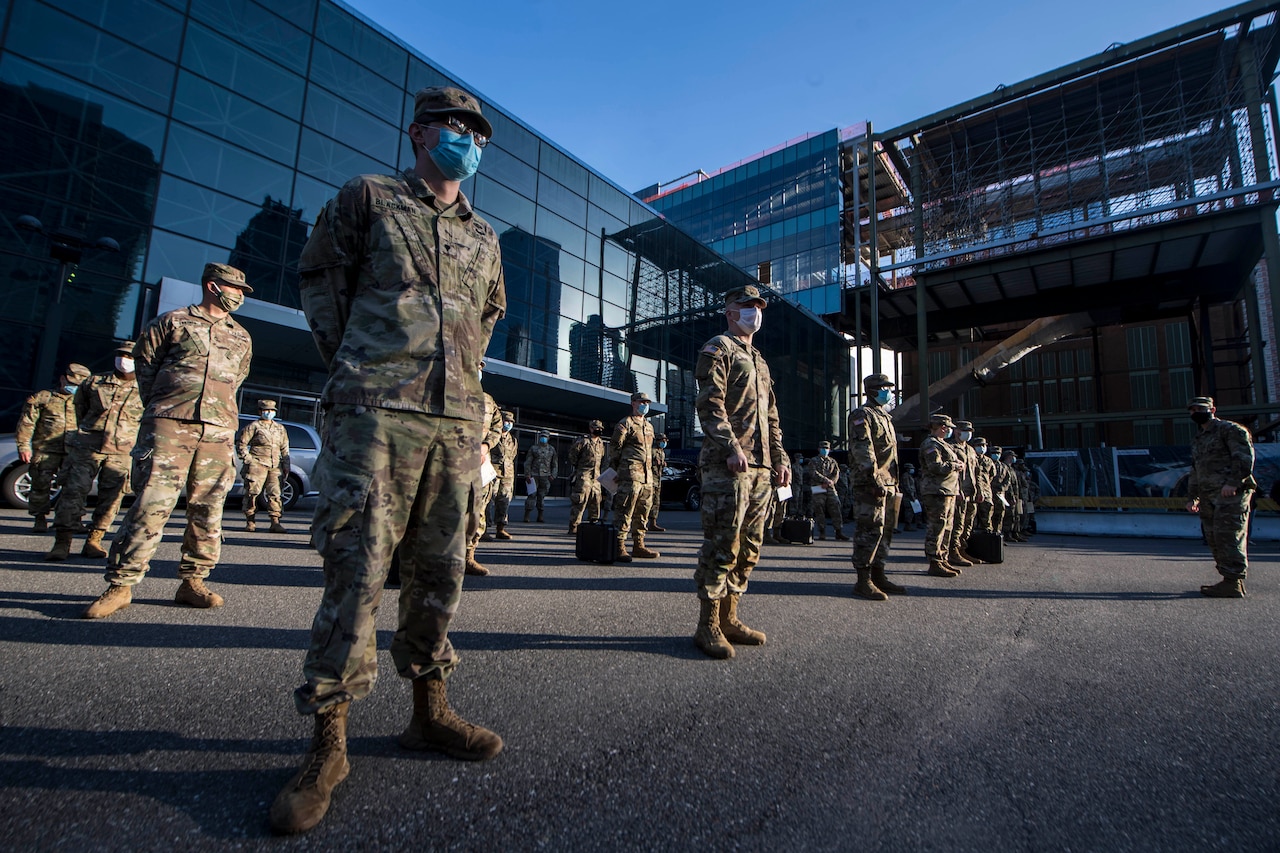 Service members wearing camouflage uniforms and face masks stand in a socially distanced formation.