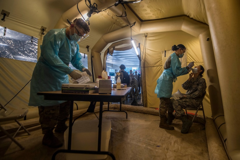 One soldier places a specimen in a bag at a table in a tent while another soldier administers a COVID-19 test to someone seated in a chair. Both soldiers are wearing personal protective equipment.