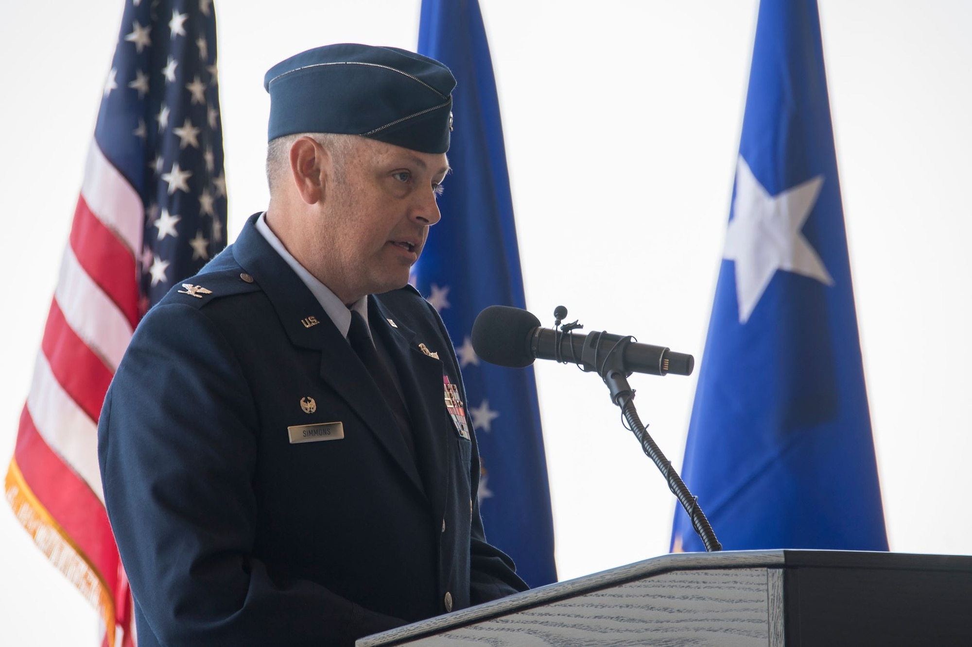 A man in dress uniform stands in front of a podium. Flags are behind him.