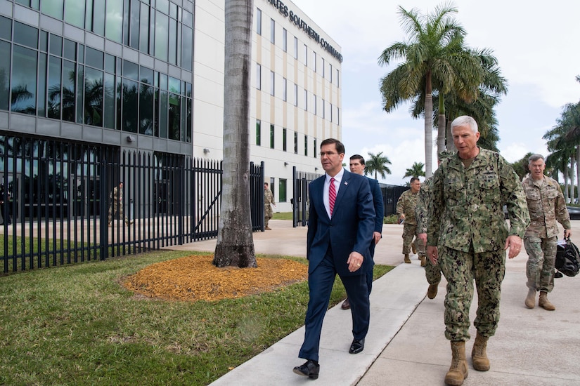 Two men, one in a Navy uniform, walk on the sidewalk past a building.