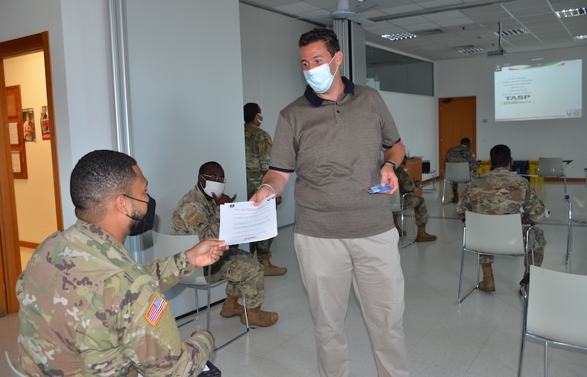 A man wearing a face mask hands pamphlets to a masked soldier seated in a classroom setting.