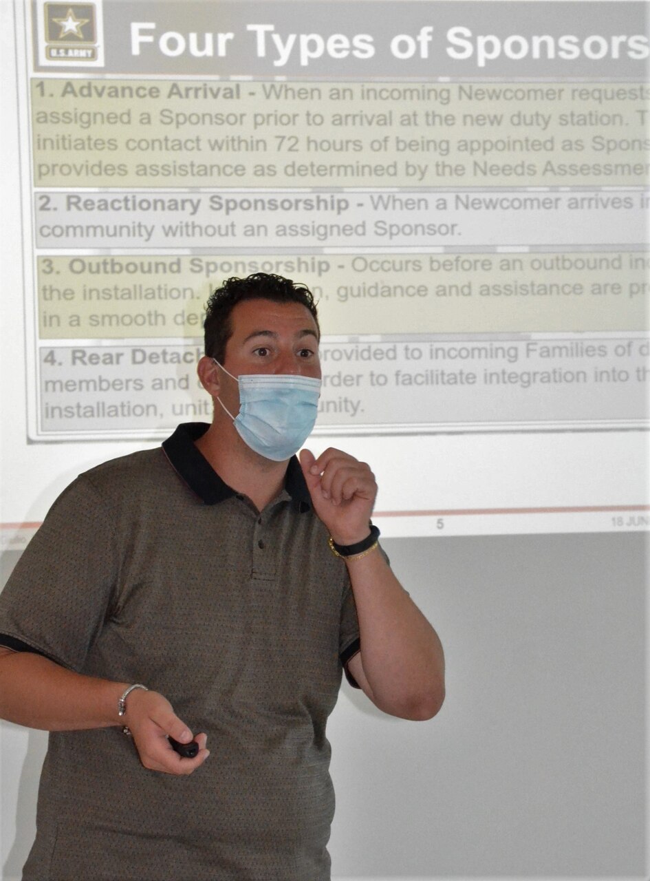 A man wearing a face mask speaks to an audience, with a briefing slide projected behind him.