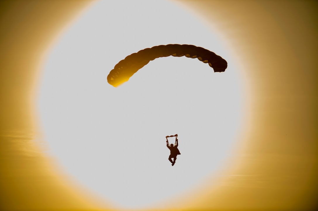 A soldier, shown in silhouette against an orange sky, parachutes with the sun in the background.