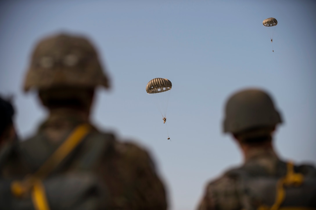 Soldiers watch fellow soldiers freefall with parachutes.