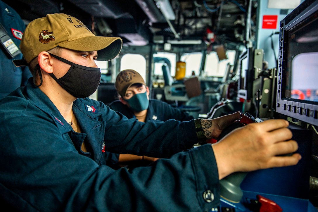 A sailor wearing protective gear holds a remote console while sitting next to another service member.