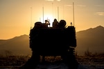 Marines are perched atop an amphibious assault vehicle, shown in silhouette, with mountains and an orange sky as the backdrop.