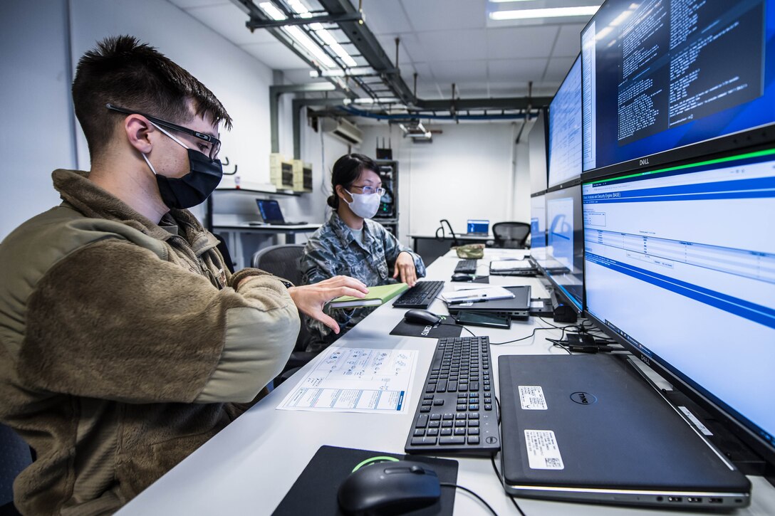 Two people sit at a table with their laptops; they stare at a wall with multiple computer monitors.