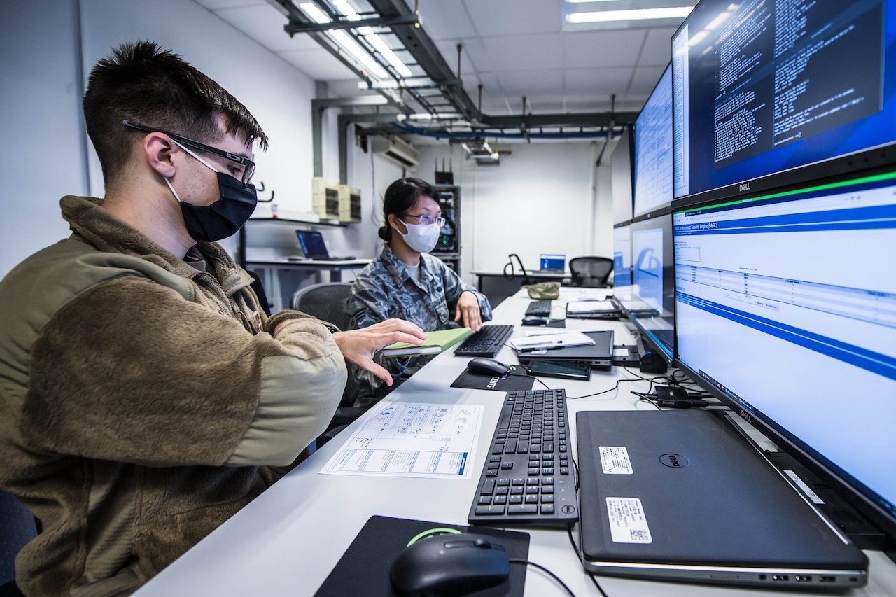 Two people sit at a table with their laptops; they stare at a wall with multiple computer monitors.