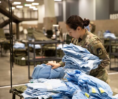 Female soldier in camo uniform sorts through blue hospital gowns.