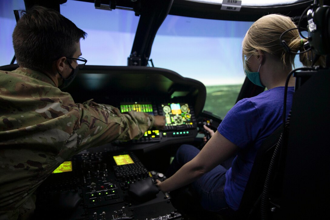 A service member and a civilian sit in a cockpit illuminated by green lights.