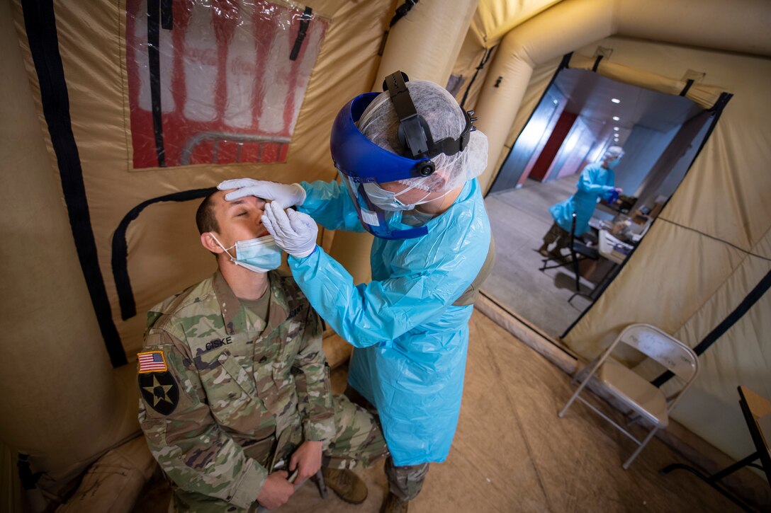 Person wearing a blue hospital gown, gloves, face mask and face shield inserts a test swab into the nose of a male soldier in camo.