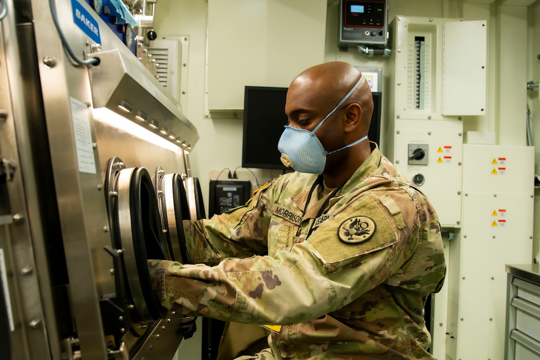 Black male soldier in camo uniform and blue surgical mask places an unseen COVID-19 test same into a machine.