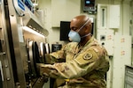 Female soldier in camo uniform sorts through blue hospital gowns.