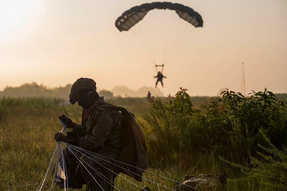A soldier kneels on the ground as another soldier descends in the sky wearing a parachute in the background.