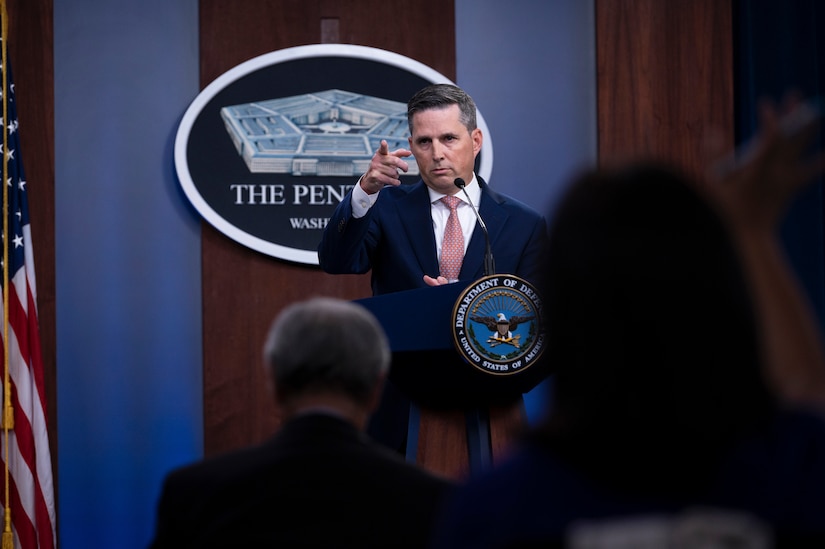 A man standing at a lectern takes questions from members of a socially distanced audience.