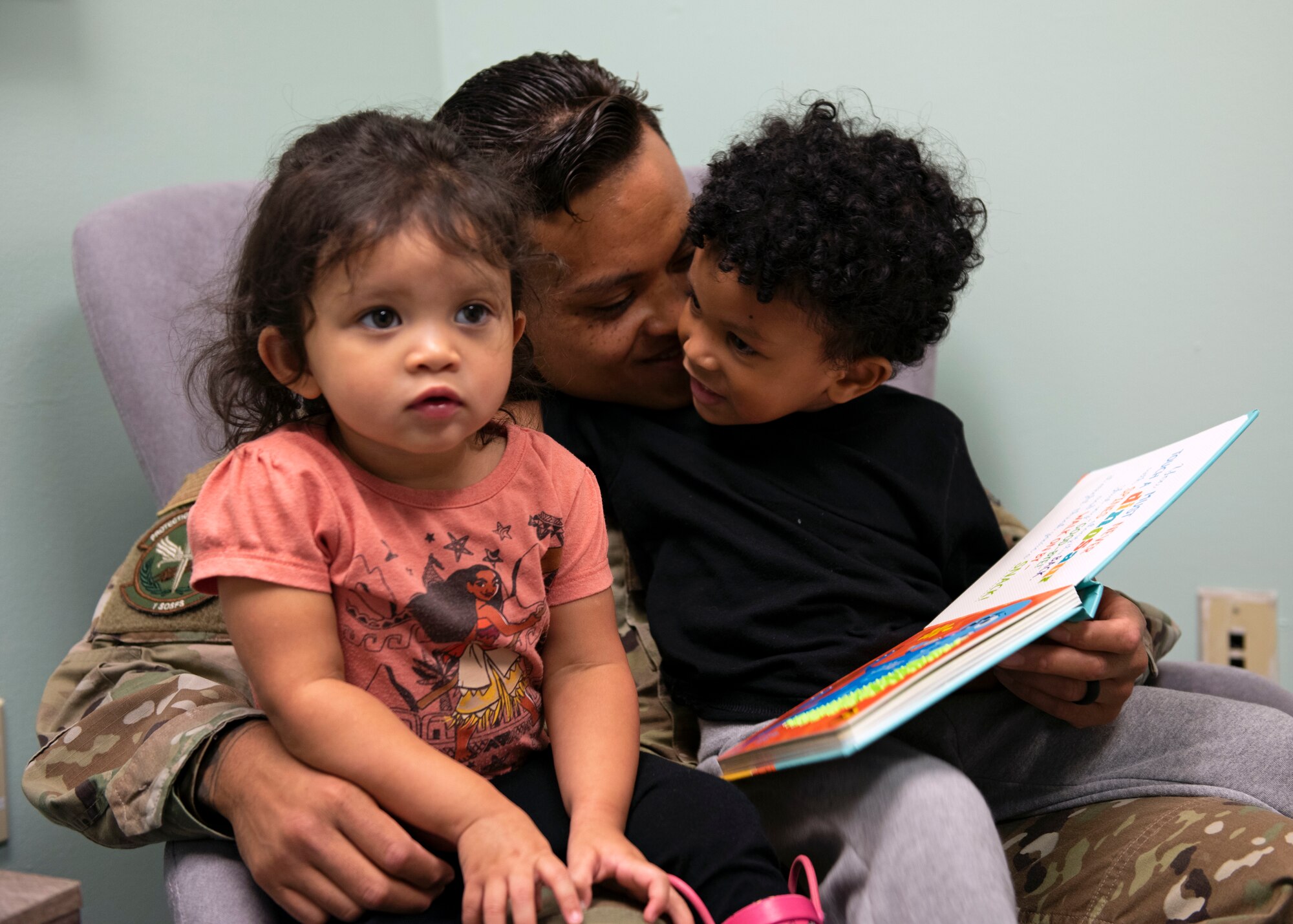 Photo of a father sitting on a chair with his two children while reading a book.