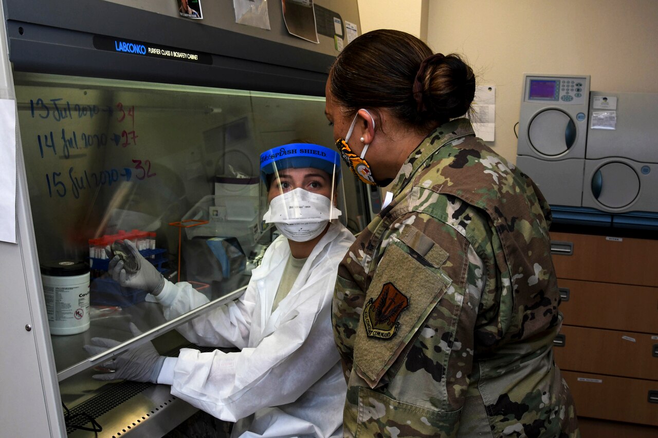 An airman stands and talks to another airman in lab gear sitting at a bench.