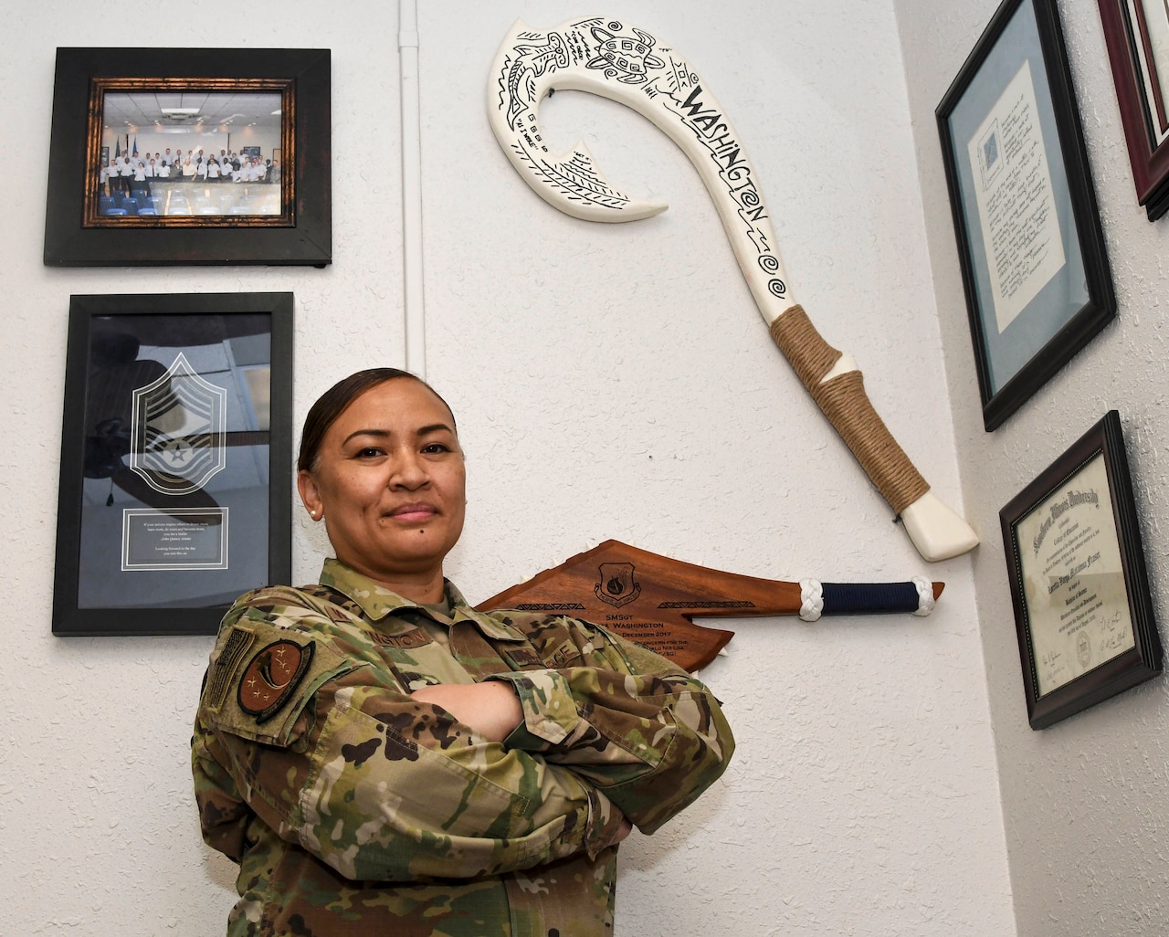 An airman stands for a photo in a room with framed items hanging on a wall.