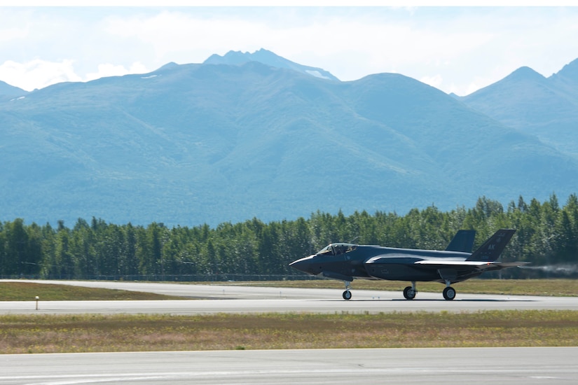 A military aircraft moves down a runway. In the background are mountains.