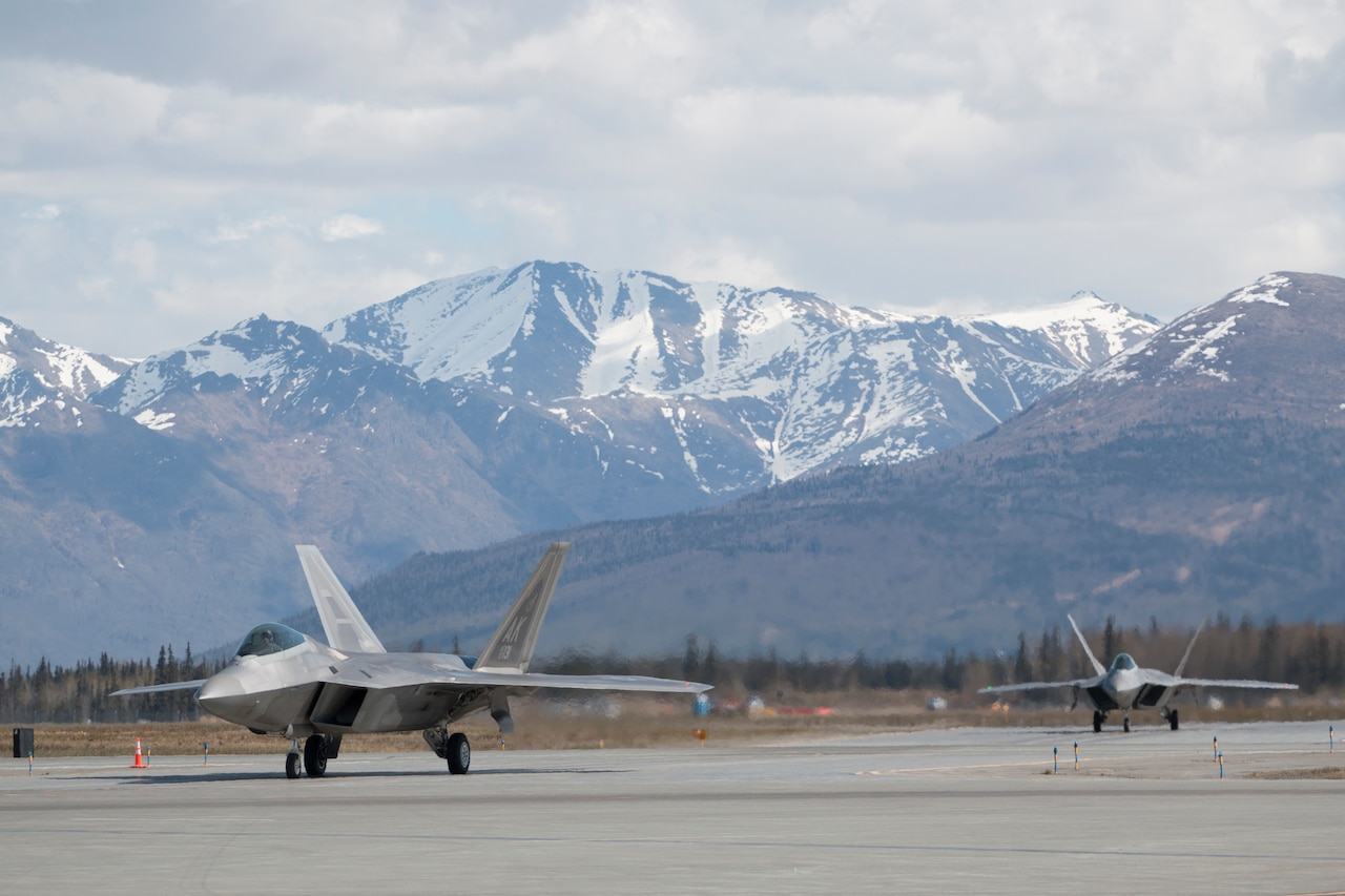 Military aircraft move down a runway. In the background are mountains.