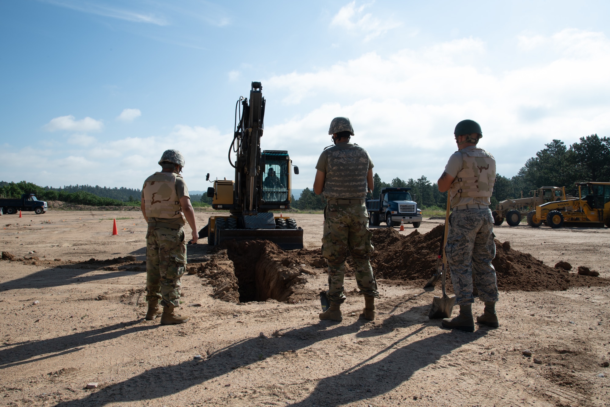 Airmen stand in front of a trench being dug by another Airman operating a heavy equipment excavator.