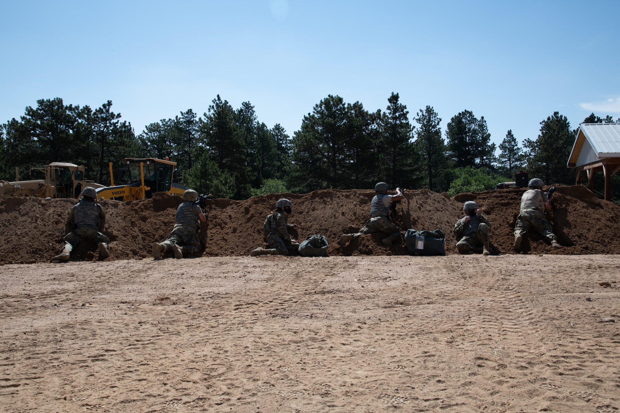 Six Airmen with rifles lined up behind a dirt mound, taking cover after hearing simulated gunfire in the distance during an exercise.