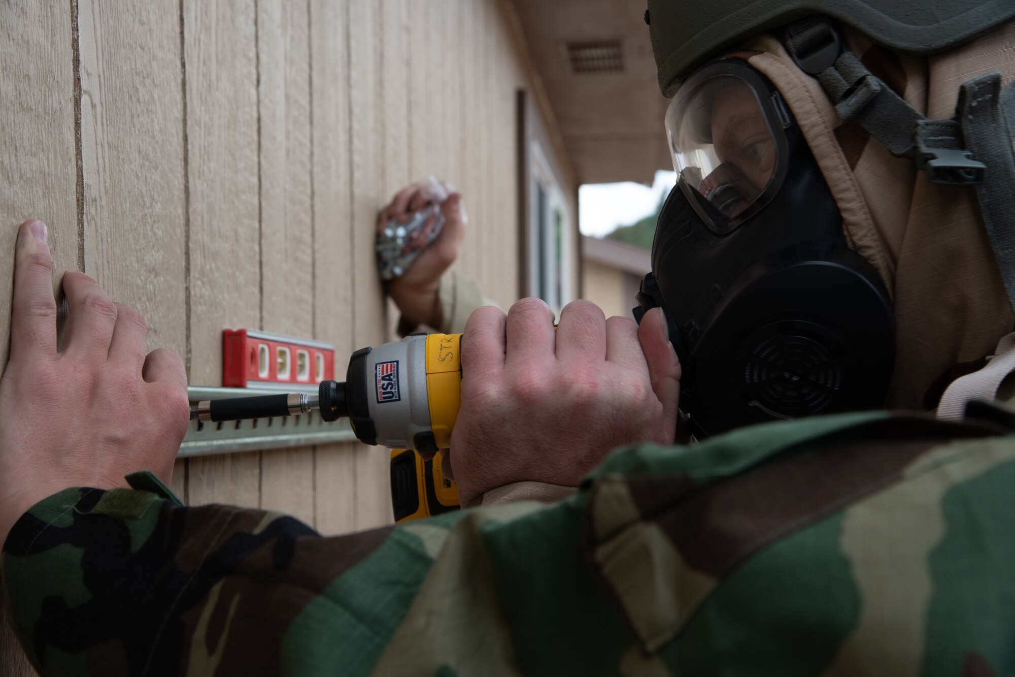 An airmen in chemical protective gear uses a power drill to mount a bracket to a wall.